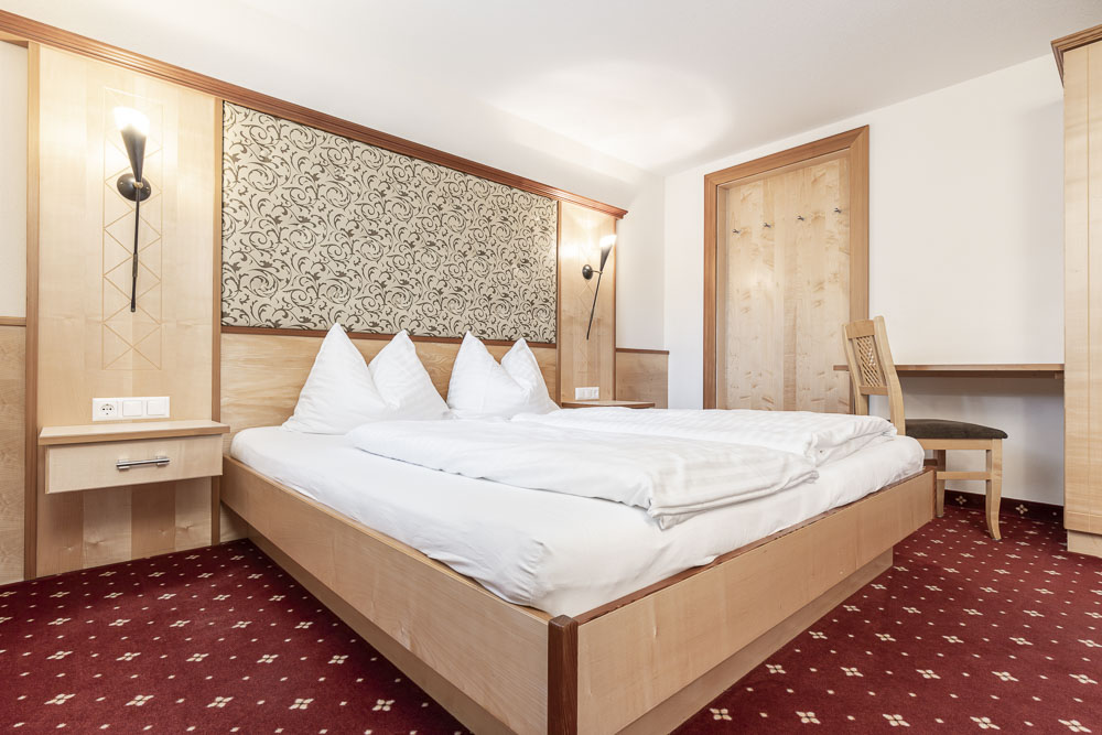 Das Alpenapart-Hotelzimmer bietet einen gemütlichen Aufenthalt mit einem großen Bett mit weißer Bettwäsche und zwei Kissen. Genießen Sie das gemusterte Kopfteil, die Holzwände und den Charme eines Teppichs mit rot-weißem Muster, perfekt für 4-6 Personen. Wandleuchten erhellen diesen einladenden Rückzugsort.
