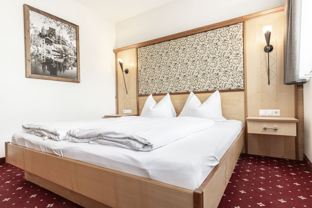Ein gemütliches Hotelzimmer im Alpenapart in Ladis, das an ein Apartment für 4-6 Personen erinnert. Es verfügt über ein großes Doppelbett mit weißer Bettwäsche und Kissen. Das Bett mit Holzrahmen verfügt über ein kunstvoll gemustertes Kopfteil, ergänzt durch Wandfotos und einen roten Teppich mit kleinen Mustern.