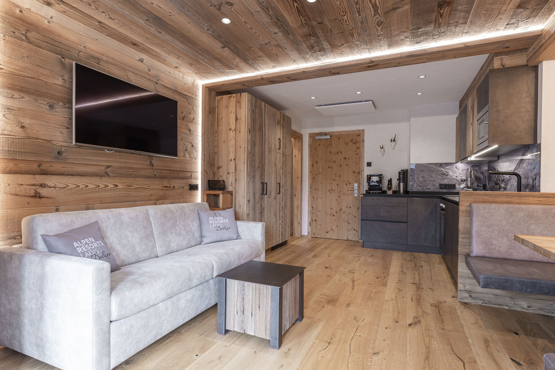Moderner Wohnraum mit Holzwänden und -boden, einem grauen Sofa, einem an der Wand montierten Fernseher und einem kleinen Couchtisch. Der Küchenbereich umfasst dunkle Schränke, eine Spüle und Geräte. Auf der rechten Seite befindet sich eine eingebaute Bank mit einem Esstisch.