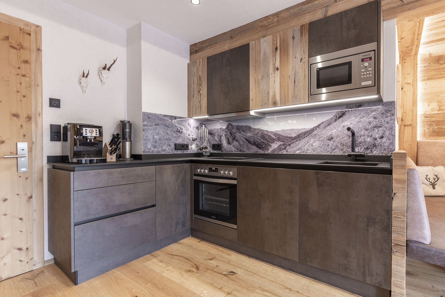 Moderne Küche mit dunklen Holzschränken und einer eleganten Arbeitsplatte. Zur Ausstattung gehören eine eingebaute Mikrowelle, ein Backofen, eine Kaffeemaschine und eine Küchenrückwand mit Berglandschaft. Helle Beleuchtung und Holzakzente sorgen für eine gemütliche Atmosphäre.