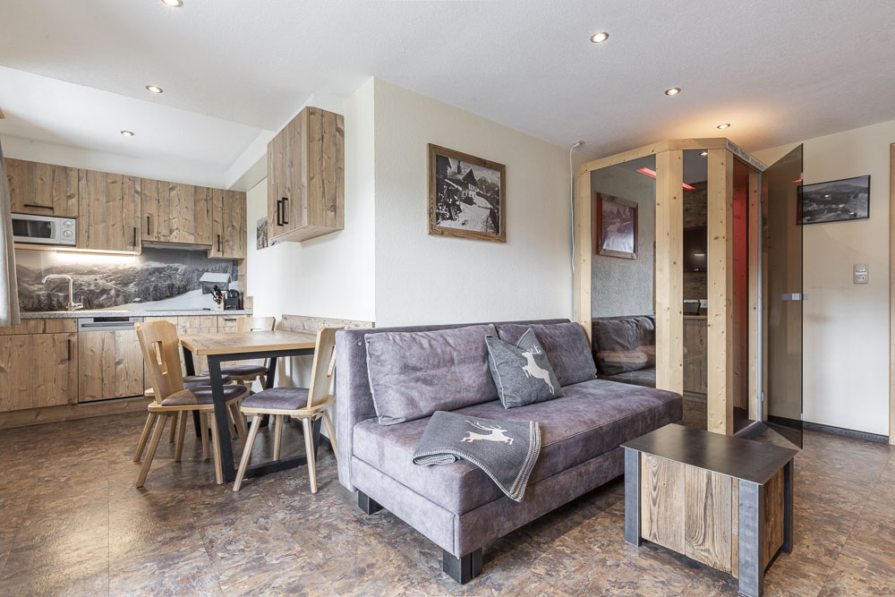 Gemütliches Apartment für 2-3 Personen in Ladis mit rustikalem Design mit Holzschränken in der Küche, einem Holzesstisch und einer grauen Couch mit einer Decke mit Hirschmuster. Warmes Licht und Kunstwerke mit Naturmotiven schmücken die Wände und schaffen eine einladende Atmosphäre.