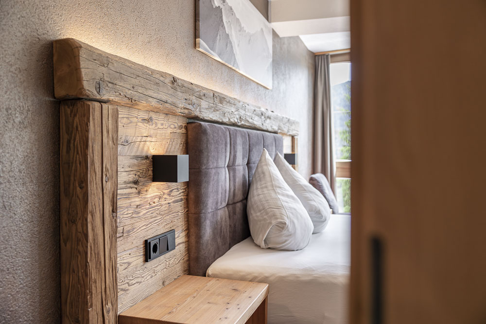 Ein gemütliches Schlafzimmer im Apartment 3 für 2-3 Personen in Ladis verfügt über ein rustikales Kopfteil aus Holz und strukturierte graue Wände. Das Bett ist hübsch mit weißen und grauen Kissen bezogen, daneben steht ein Nachttisch aus Naturholz mit einer schwarzen Lampe. Licht strömt durch ein teilweise sichtbares Fenster herein.