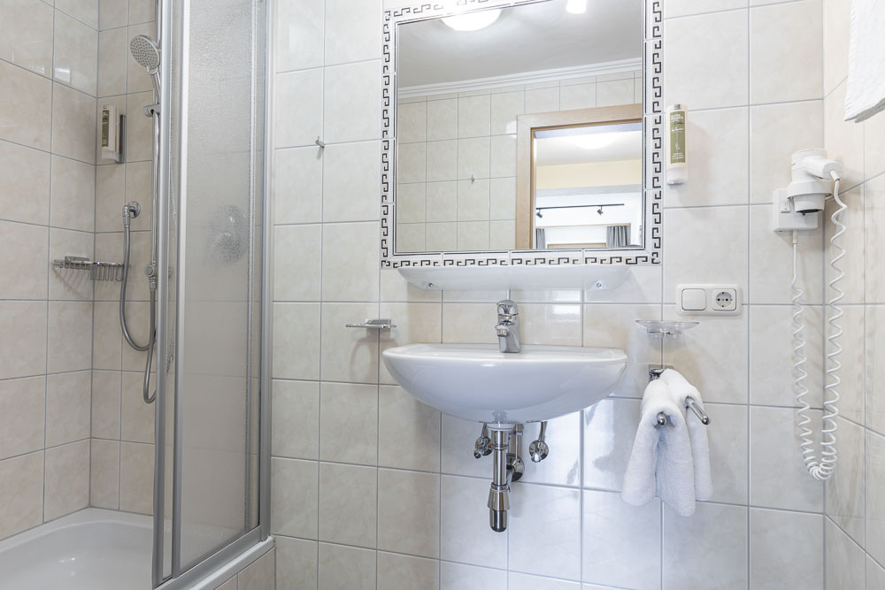 Ein kleines, weiß gefliestes Badezimmer im Apartment 4 für 2-5 Personen in Ladis verfügt über eine Duschkabine, ein wandmontiertes Waschbecken und einen rechteckigen Spiegel mit gemustertem Rand. Ein Haartrockner ist an der Wand befestigt und zwei Handtücher hängen an einem Ständer neben dem Waschbecken.