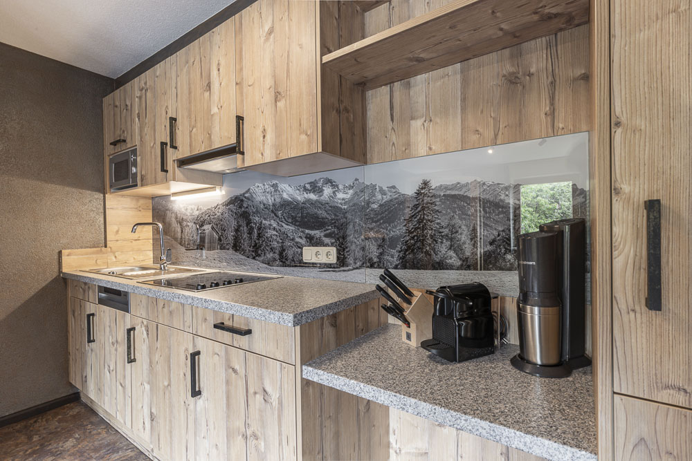 Moderne Küche im Apartment 4 für 2-5 Personen in Ladis mit Holzschränken, Granitarbeitsplatten und einer Küchenrückwand mit Berglandschaftsmotiv. Ausgestattet mit Herd, Spüle, Mikrowelle, Kaffeemaschine und einem Messerset auf der Theke.