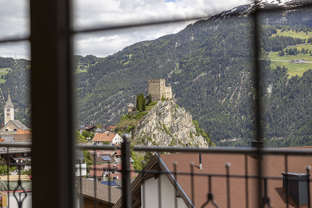 Eine steinerne Burg auf einem Hügel, durch ein Fenster betrachtet, umgeben von Bergen und den Dächern eines Dorfes.