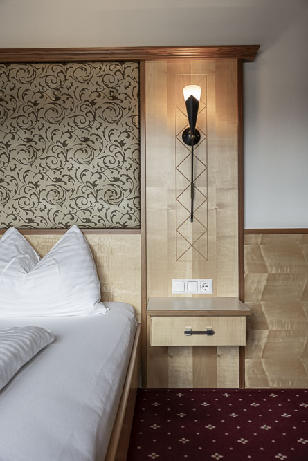 Nachttisch im Hotelzimmer mit Wandlampe, Steckdosen und gemustertem Kopfteil.