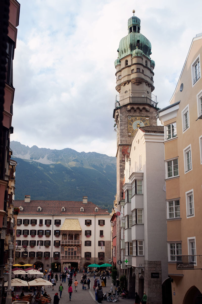 Stadtplatz mit einem hohen Uhrenturm, Menschen, die spazieren gehen, und Bergen im Hintergrund.