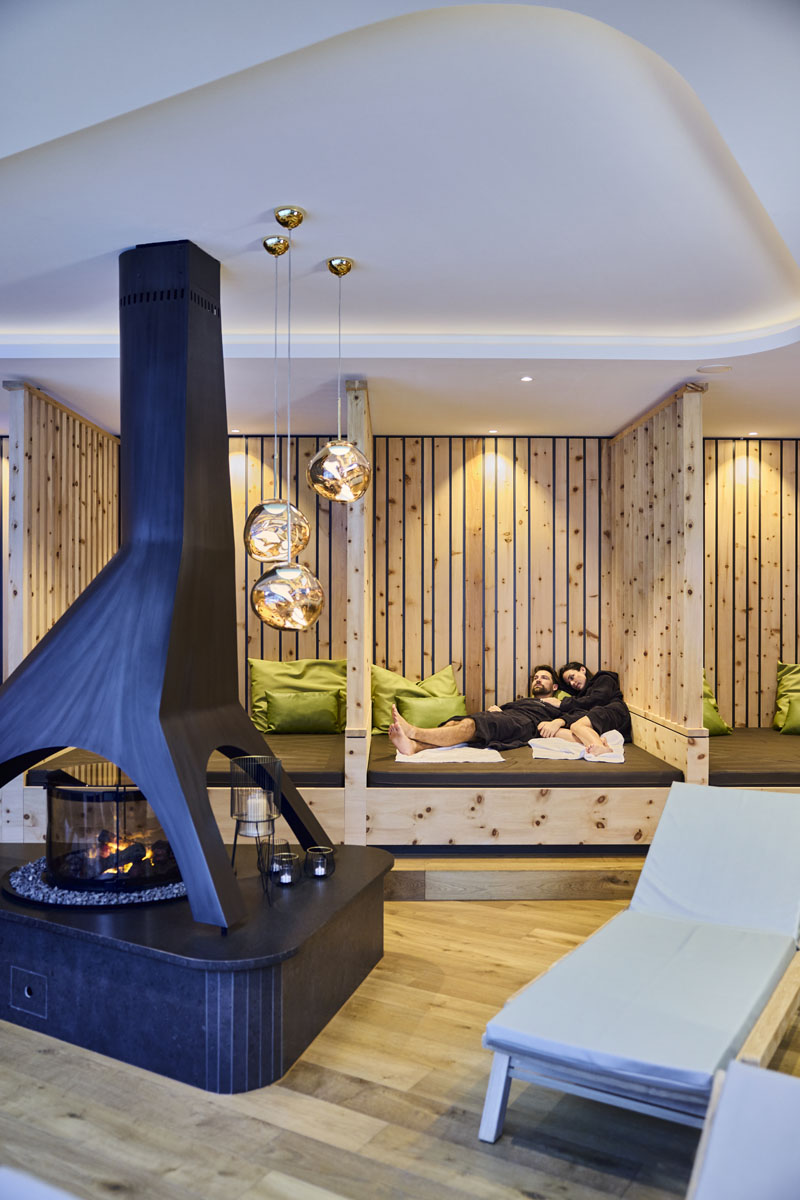 Zwei Personen entspannen sich auf einer Holzbank vor einem modernen schwarzen Kamin in einem gemütlichen Lounge-Bereich.