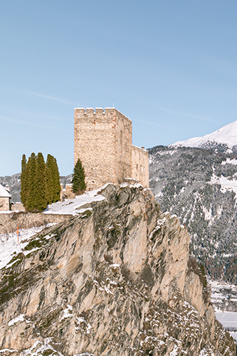 Steinschloss auf einer schneebedeckten Klippe, mit Blick auf Alpenresorts und Berge im Hintergrund.