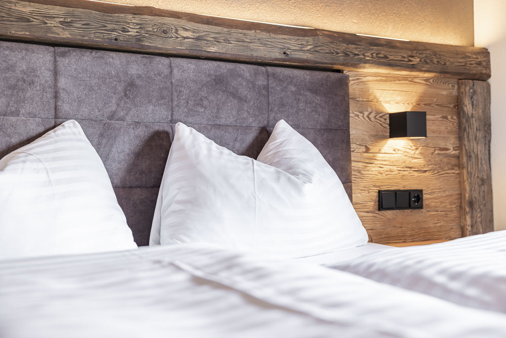 Nahaufnahme eines ordentlich gemachten Bettes mit weißen Kissen und hölzernem Kopfteil, warm beleuchtet.