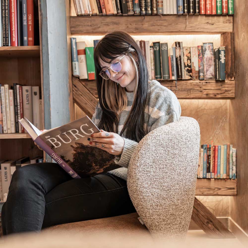 Frau mit Brille liest BURG in einer gemütlichen Bibliothek in Mimis Kinderspielwelt im Alpenapart in Ladis.