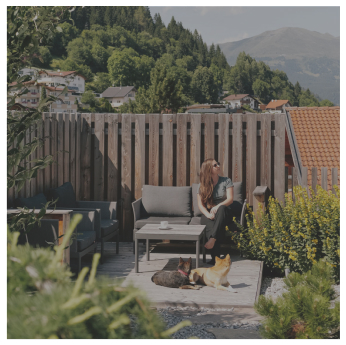 Frau auf einer Veranda mit zwei Hunden, Berge, Häuser und Alpenresorts im Hintergrund.